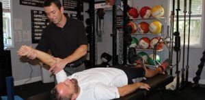 A physical therapist specializing in manual therapy Bergen County NJ assists a man lying on a table by stretching his arm in a gym, with colorful medicine balls and workout equipment visible in the background.