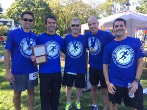 Five men stand outdoors in a park, smiling and wearing matching blue "The Recovery Room" shirts—celebrating injury prevention Bergen County NJ. Two display medals, one holds a plaque, and another has a water bottle. Trees and a tent complete the scene.