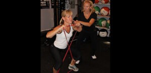 Two women exercise in a gym; one performs a resistance band squat while the other, likely a physical therapist Bergen County NJ, offers support, with medicine balls stacked on shelves in the background.