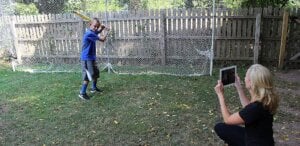 A woman kneels on grass, holding a tablet to record a young boy with a bat who is ready to swing in a backyard enclosed by netting and a wooden fence—capturing moments physical therapists Bergen County NJ value for assessing movement skills.