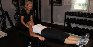 A trainer, possibly a physical therapist Bergen County NJ, assists a woman lying on her back on a gym bench, gently supporting her head. The room has free weights and gym equipment as sunlight streams in through the window.